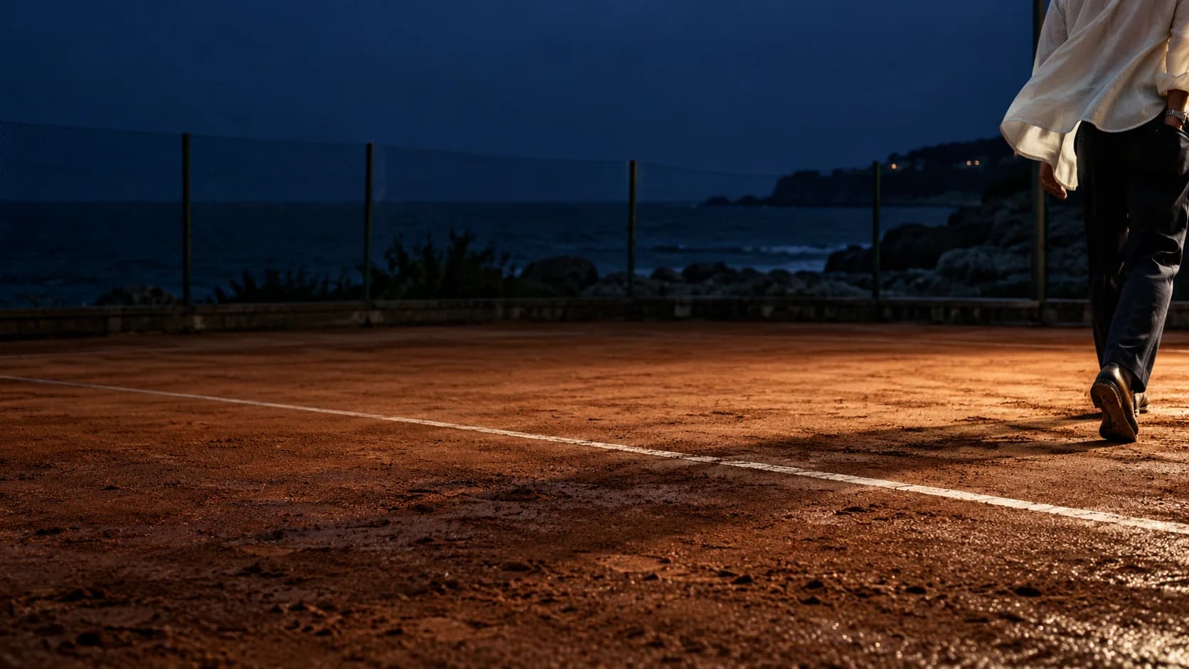 A figure leaving a seaside tennis court at blue hour, ivory fabric catching the light above the worn clay.
