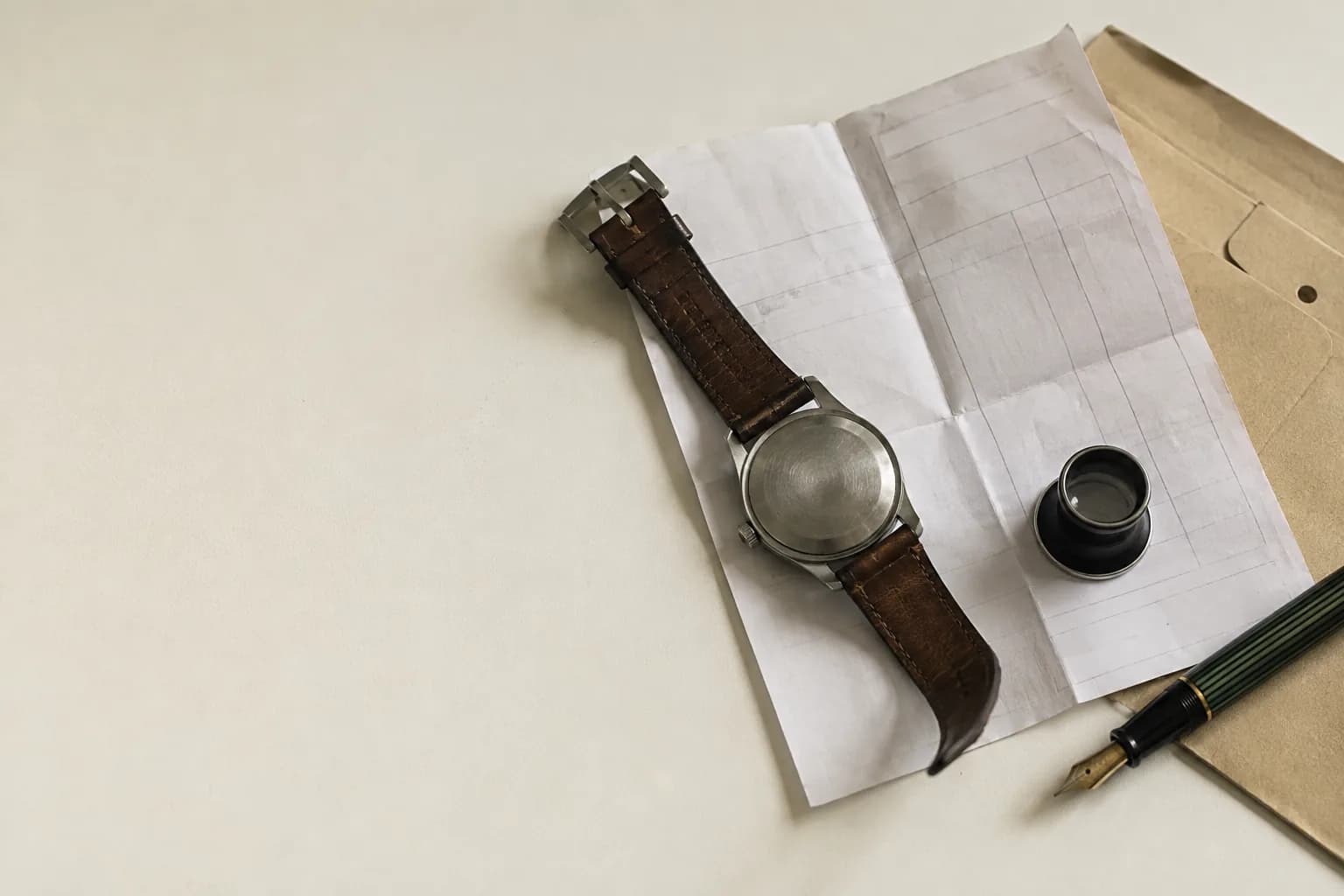 A wristwatch lying face down on a folded service invoice, beside a loupe, a document sleeve, and a fountain pen.