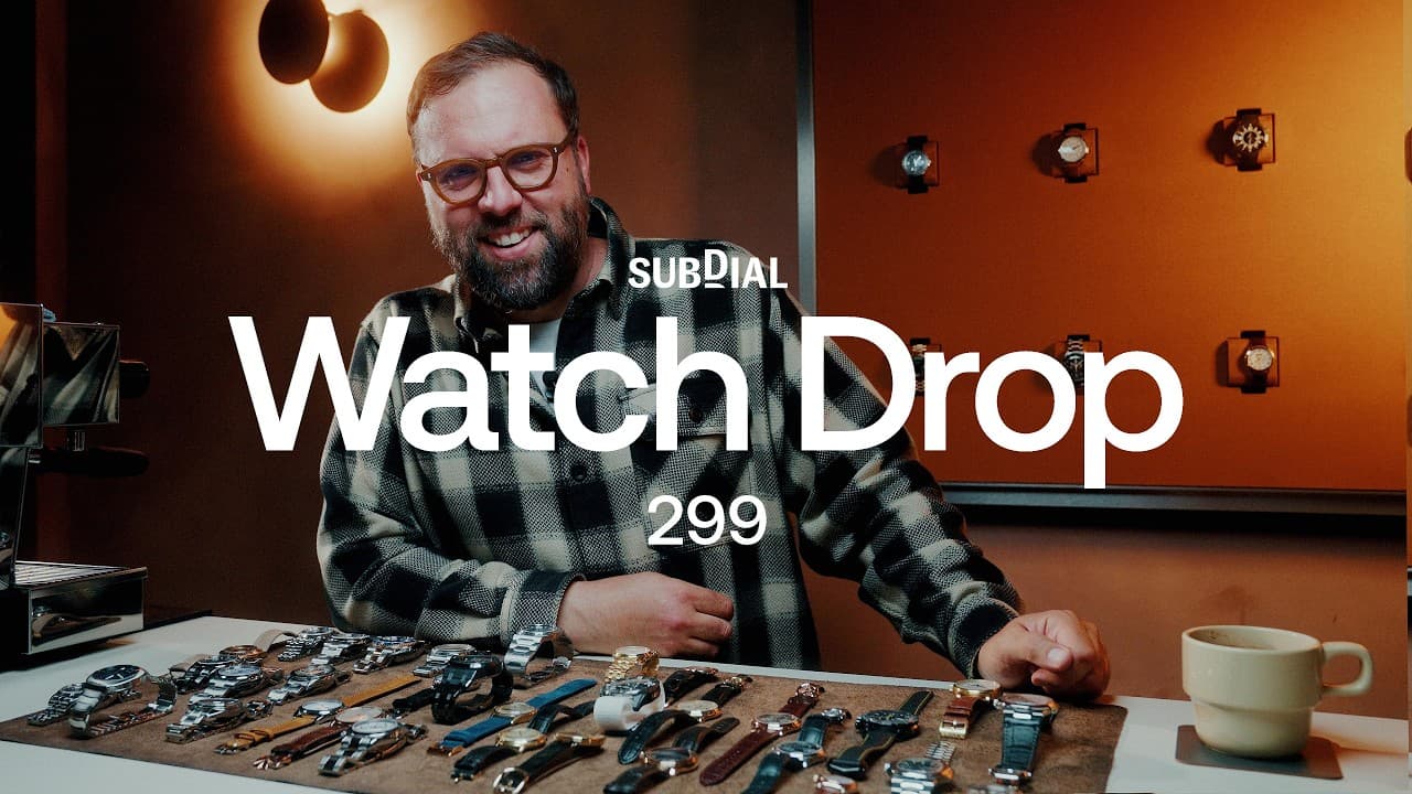 A smiling man stands behind a table covered with many wristwatches in a warmly lit room.