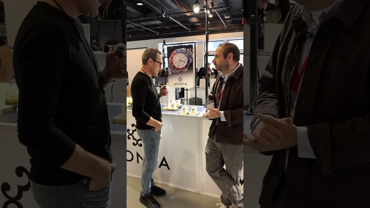 Two men talk beside a watch display booth with a large wall sign and several watches under glass.