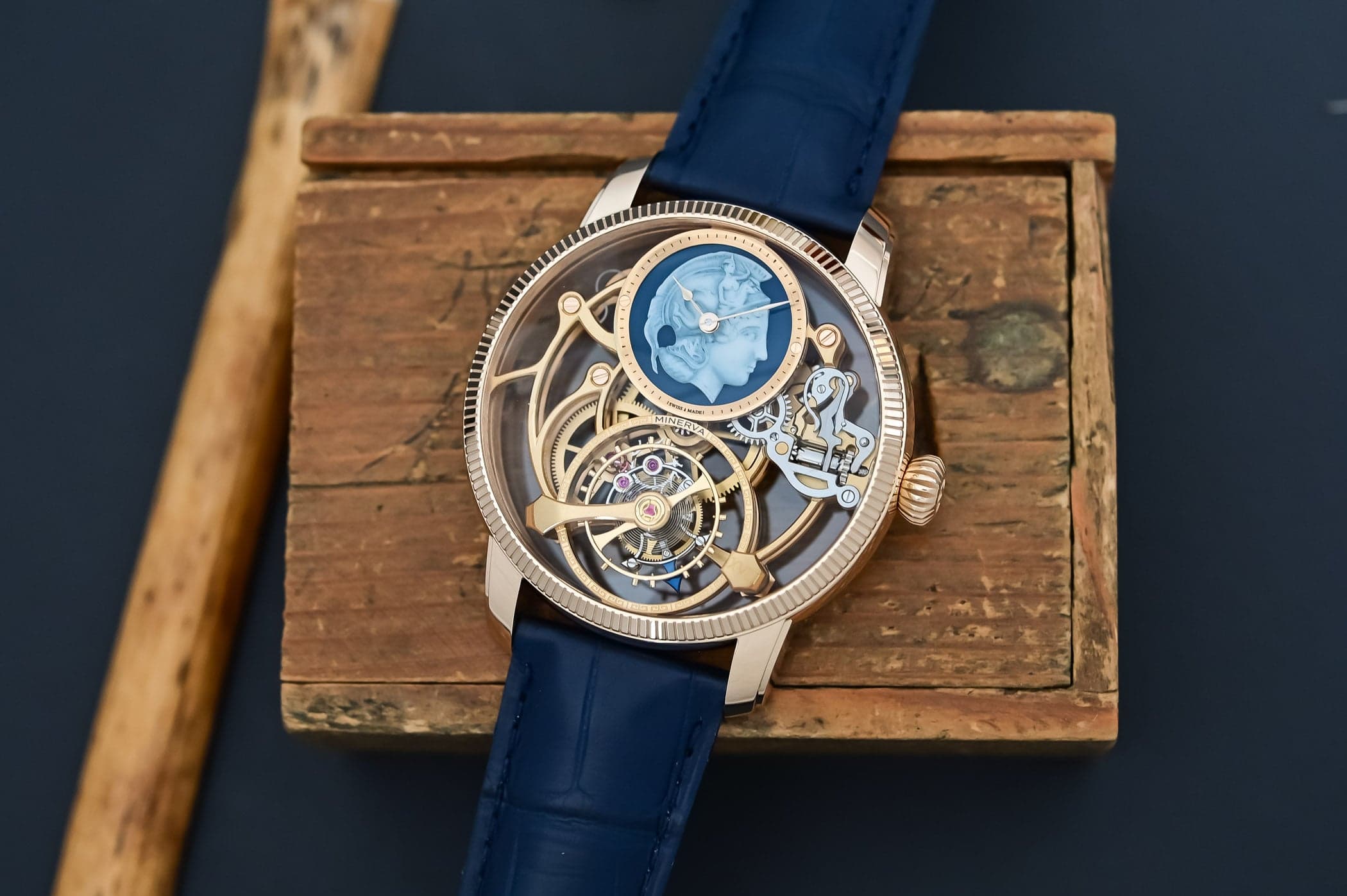 A skeletonized wristwatch with a blue strap rests on a wooden box, showing a large exposed movement and a blue medallion at the top of the dial.