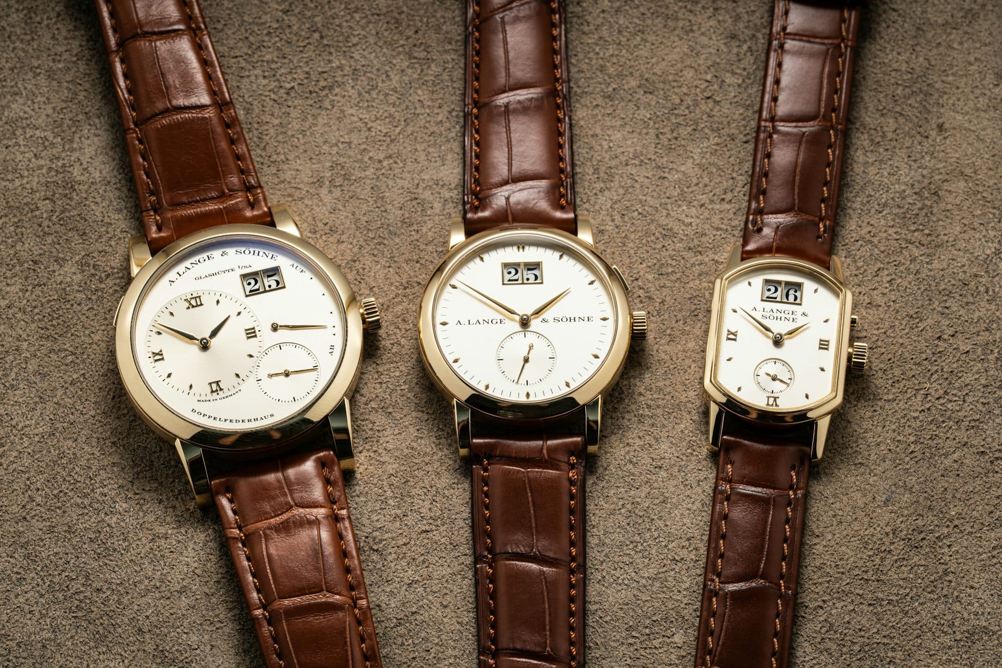Three gold wristwatches with brown leather straps lie side by side on a textured surface.
